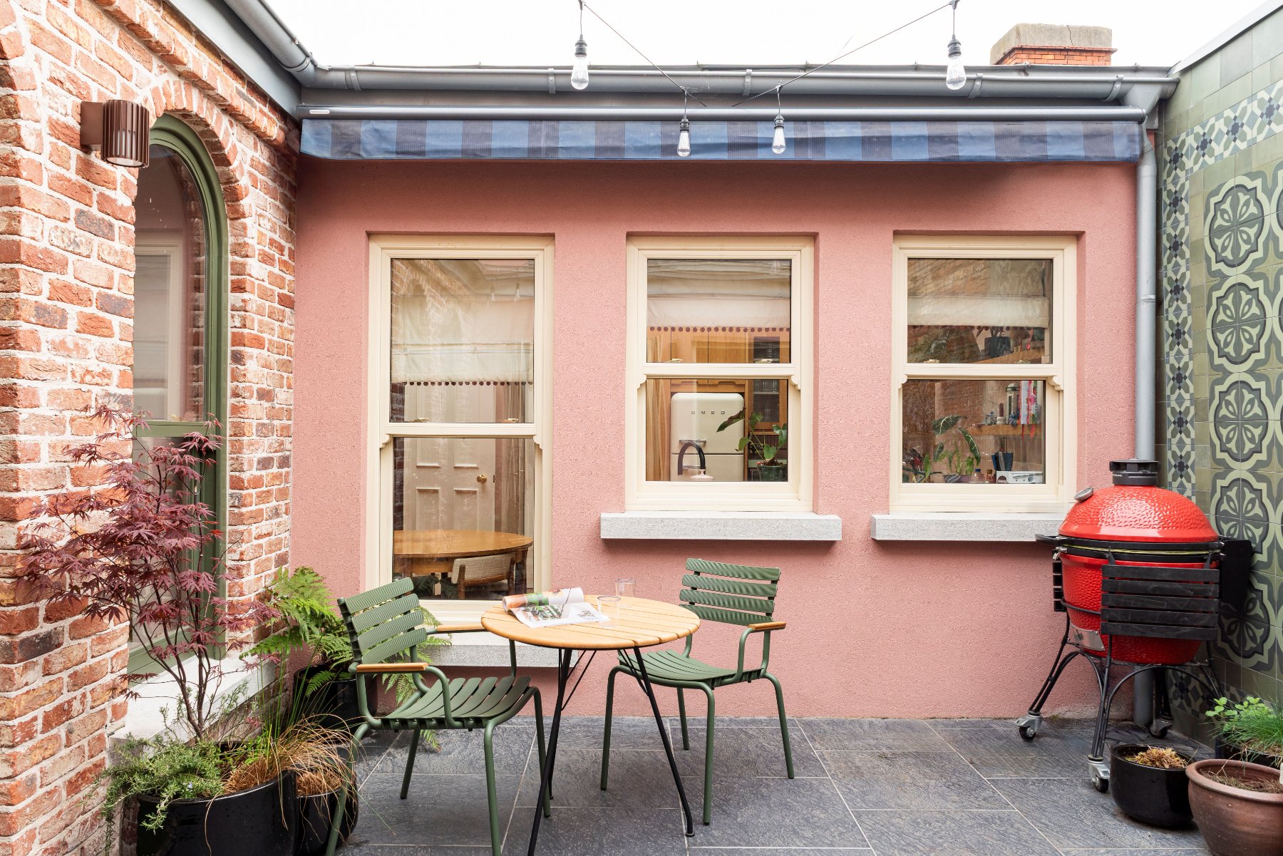 The courtyard: a bistro table and green chairs against the pink wall, with festoon lights, a red kamado grill and patterned tiles